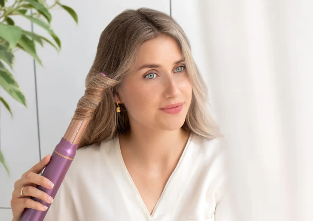 Kobieta trzyma różową lokówkę z cylindryczną złotą nasadką i nawija pasmo blond włosów, stojąc w jasnym pomieszczeniu.
