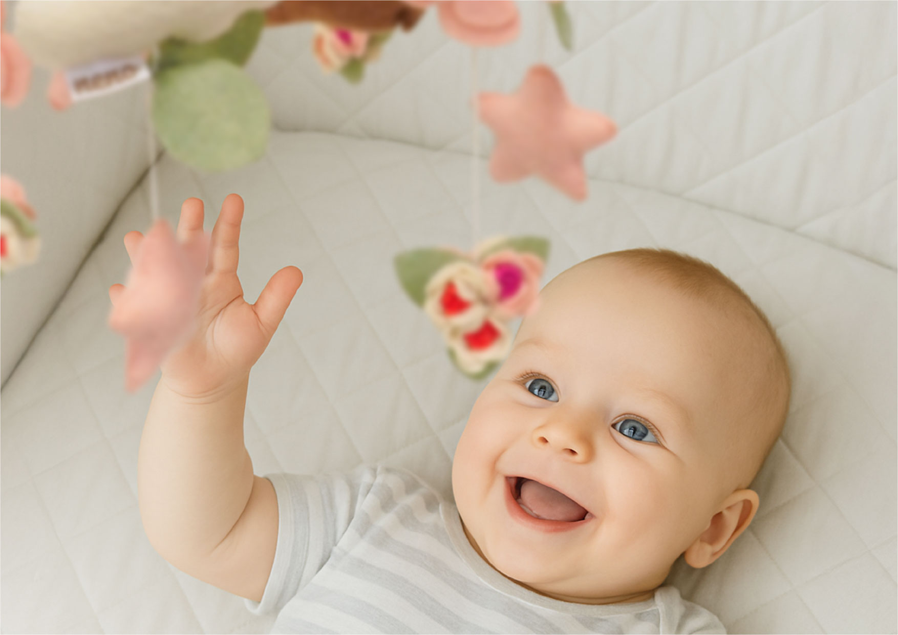 Uśmiechnięte dziecko wyciąga rączkę do maskotek karuzeli Neno Rosi; zawieszki zachęcają do pierwszych prób chwytania i koordynacji.